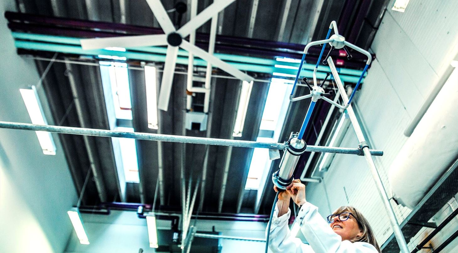 Laborant i hvid kittel og briller mler luftstrm med professionelt mleudstyr i et ventilationssystem med synlige kanaler og rr i loftet